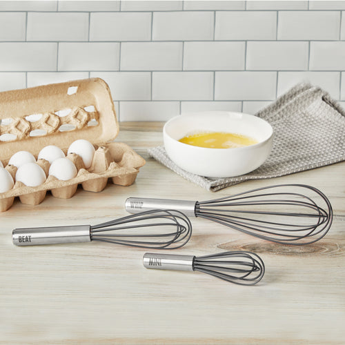 Three stainless steel Beat Whisks with metal handles labeled “BEAT,” “WHIP,” and “MIX” rest on a light wood surface alongside a carton of eggs, a bowl of beaten eggs, and a gray cloth. These dishwasher-safe kitchen essentials are must-haves.