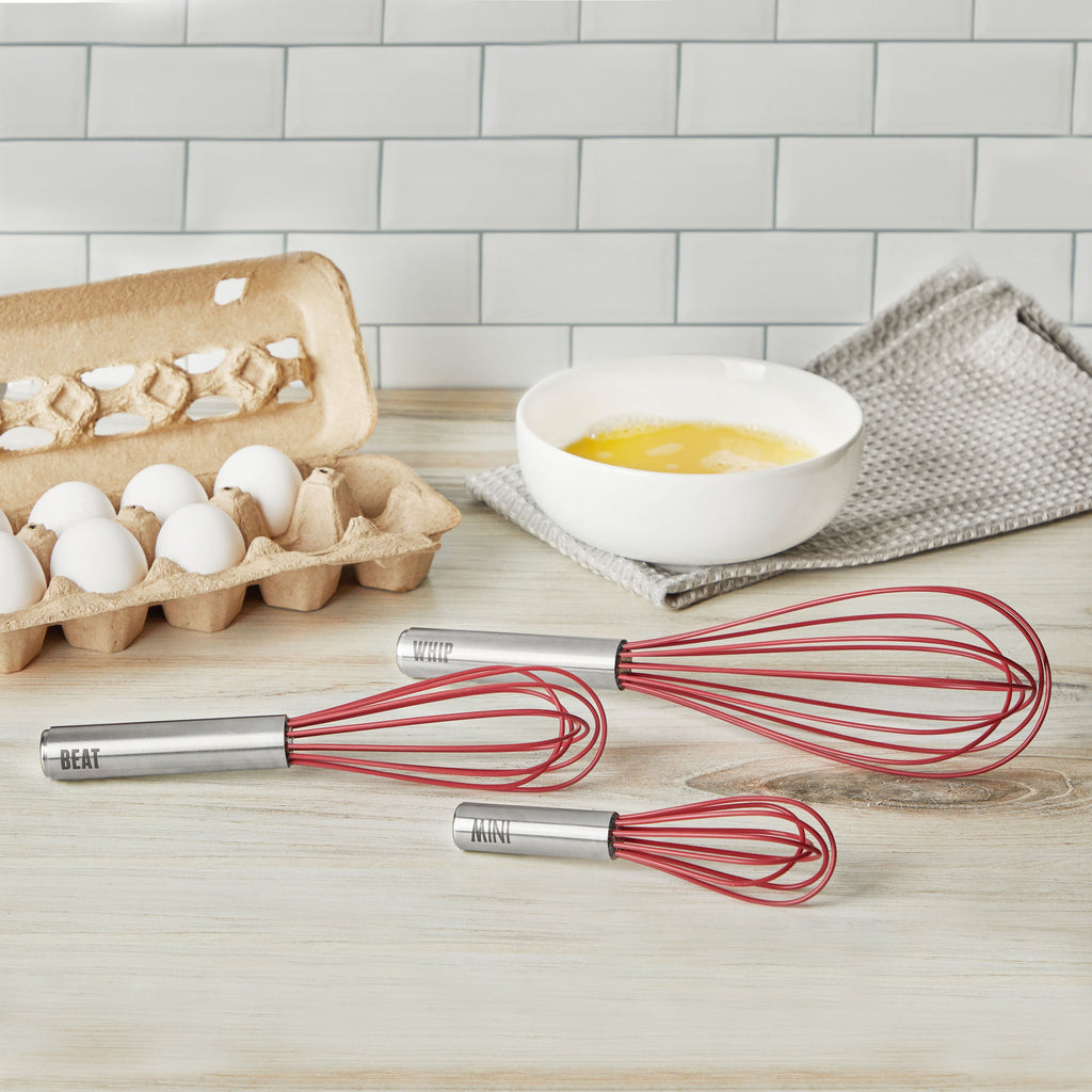 Three red Whip Whisk balloon whisks with stainless steel handles labeled BEAT, WHIP, and MIX rest on a light wood countertop beside eggs, a bowl of beaten eggs, and a gray kitchen towel. White tile backsplash in the background.