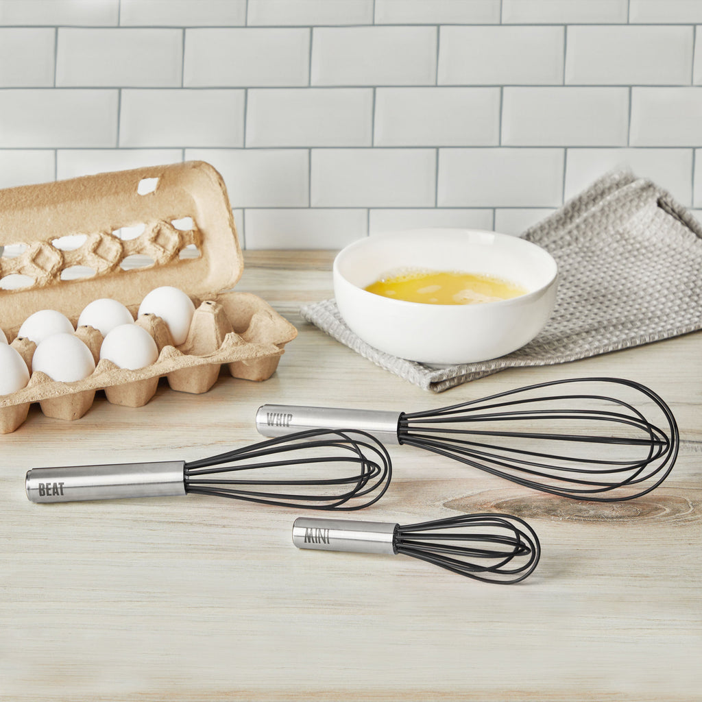 The Mini Whisk, along with two black-handled whisks labeled BEAT, WHIP, and MIX, sit on a counter next to a carton of eggs and a white bowl of beaten eggs, with a gray towel and tile backsplash in the background.