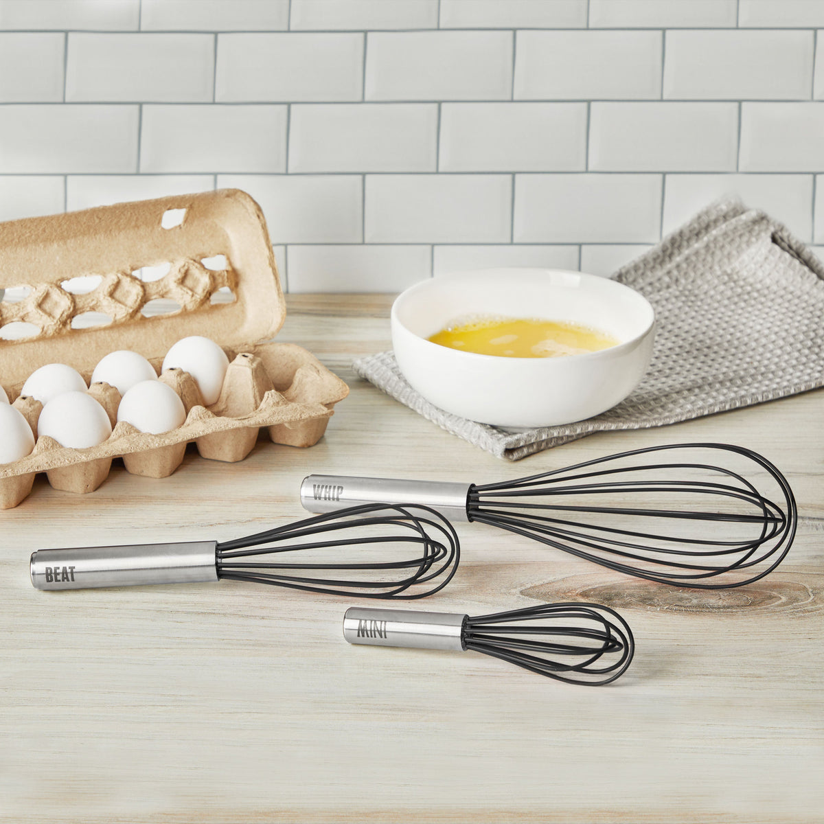 The Mini Whisk, along with two black-handled whisks labeled BEAT, WHIP, and MIX, sit on a counter next to a carton of eggs and a white bowl of beaten eggs, with a gray towel and tile backsplash in the background.