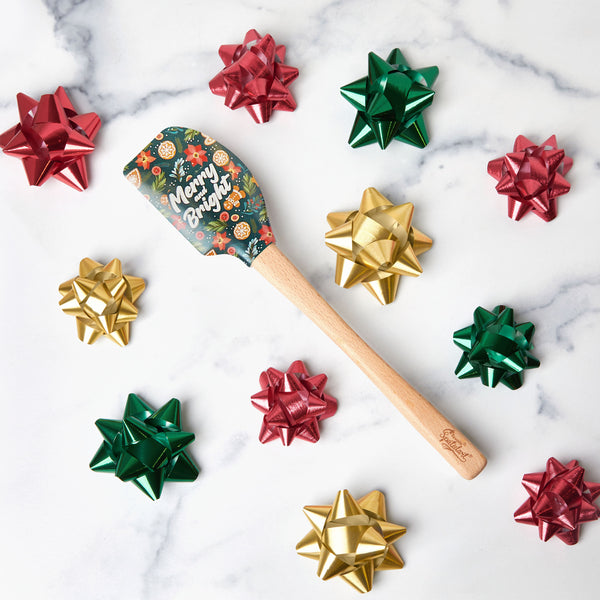 The Holiday Spatulart, featuring a wooden handle and a Merry & Bright festive pattern, is shown among shiny red, green, and gold gift bows on white marble—perfect as a gift wrap spatula for seasonal baking.