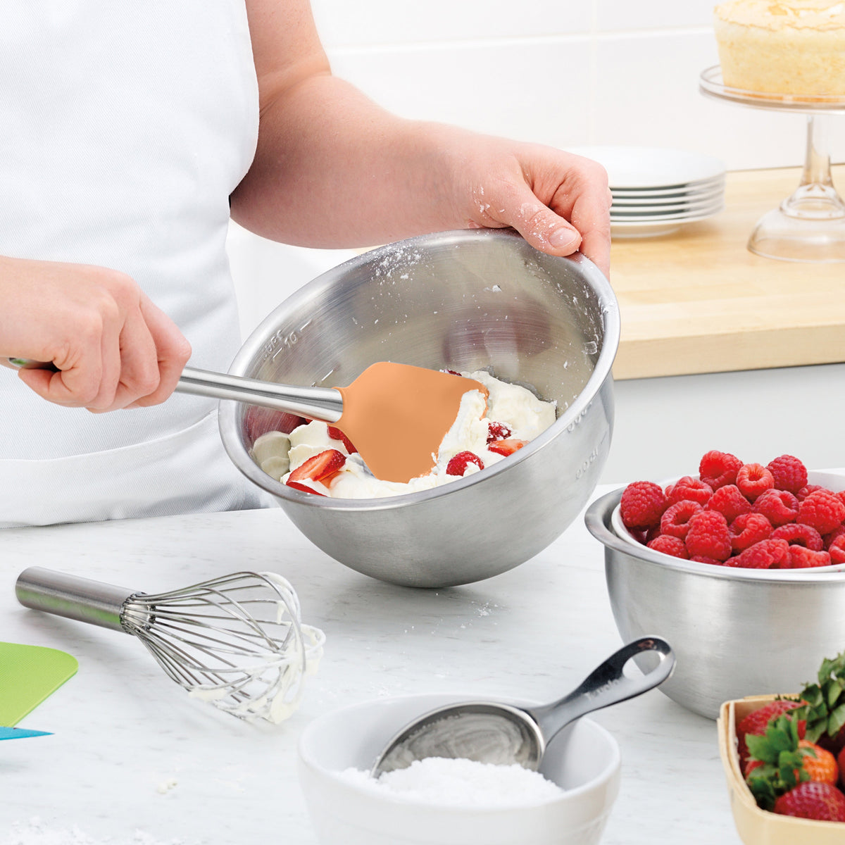 In a bright kitchen, someone in a white apron mixes cream and strawberries in a metal bowl with the durable Flex-Core Spatula, while a whisk, powdered sugar, and a bowl of raspberries sit on the counter nearby.