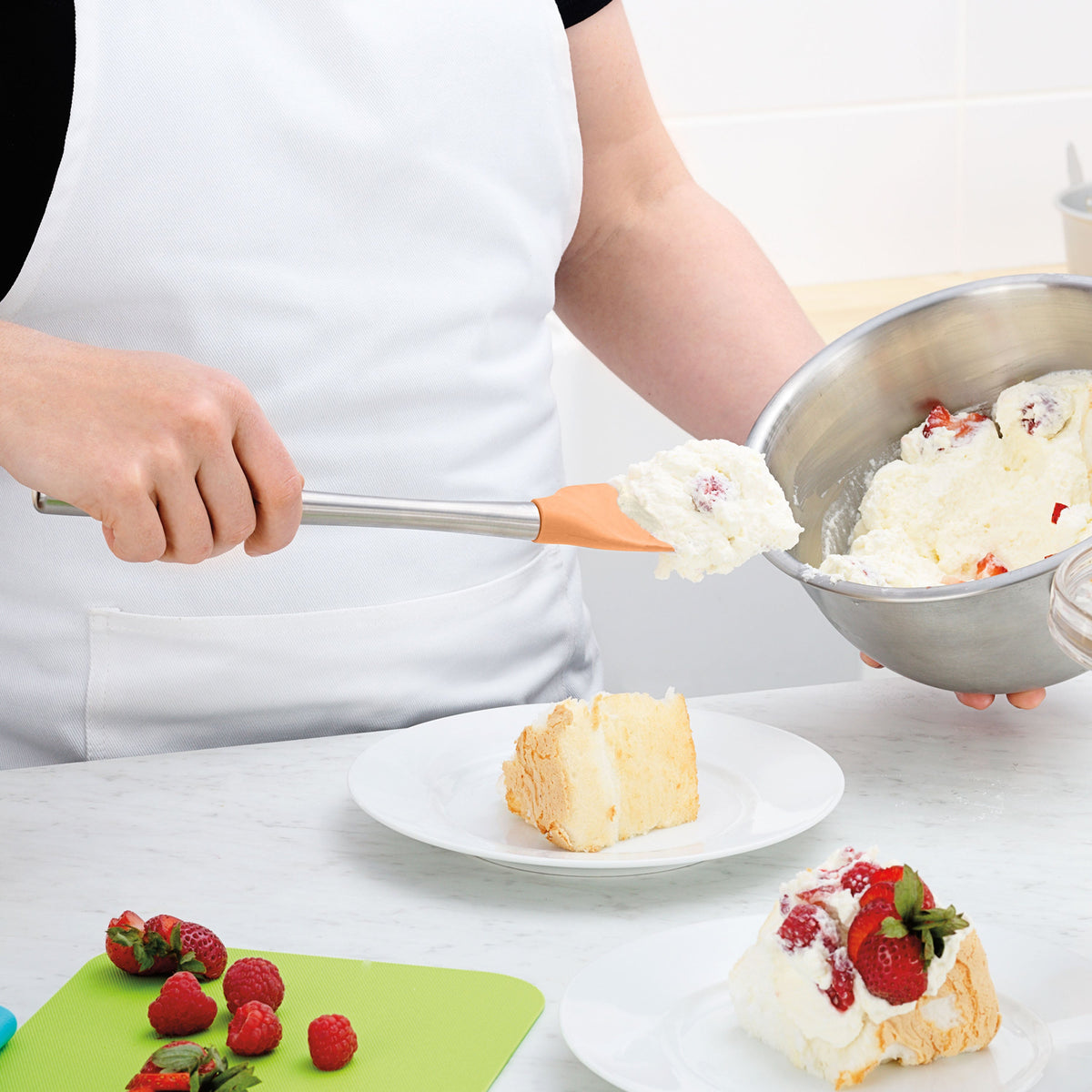 Wearing a white apron, a person uses the tovolo Flex-Core Silicone Spoonula to top cake with whipped cream. Fresh strawberries and raspberries sit on a nearby cutting board.