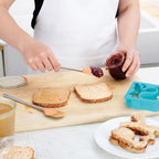 Someone uses the tovolo Flex-Core Mini Spatula + Spoonula with a stainless steel handle to spread jelly on bread near another slice with peanut butter. Shaped sandwiches and a sandwich cutter rest on a wooden cutting board.