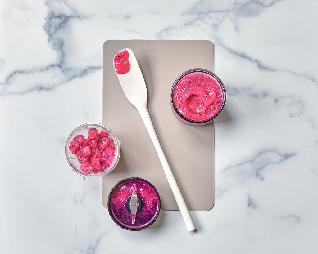 The tovolo Flex-Core Blender Spatula with a long white handle and pink smoothie on it rests on a beige board, surrounded by a jar, blender cup with chopped fruit, and blender blade containing vibrant pink smoothie, atop a marble surface.