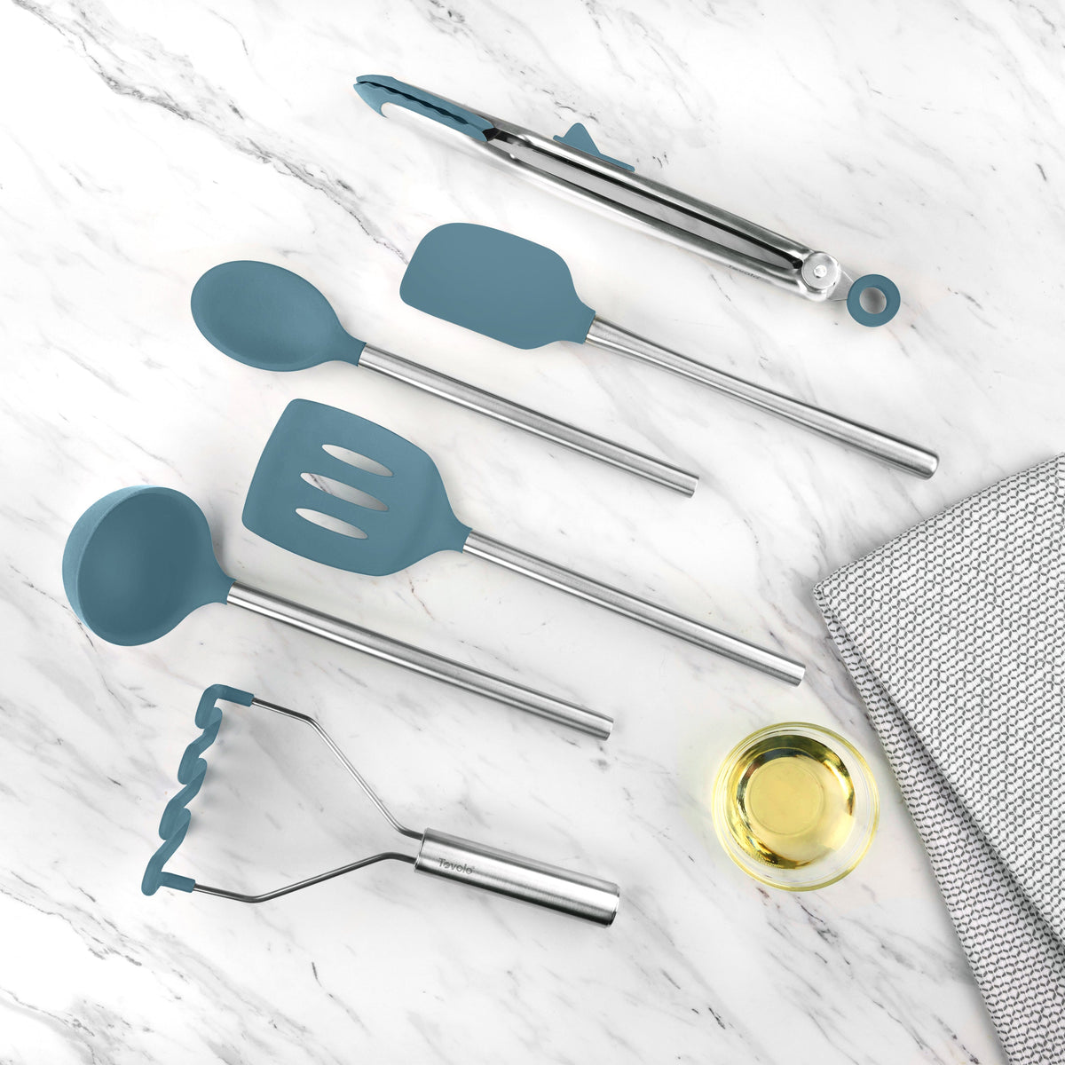 The tovolo Silicone and Stainless Steel Utensil Set of 6 is displayed on a white marble surface beside a folded gray towel and a small glass bowl filled with oil.