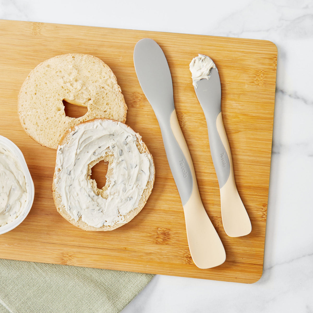 Two bagel halves with cream cheese sit on a wooden cutting board beside a tub of cream cheese and two Scoop and Spread tools, all arranged on a white marble surface.