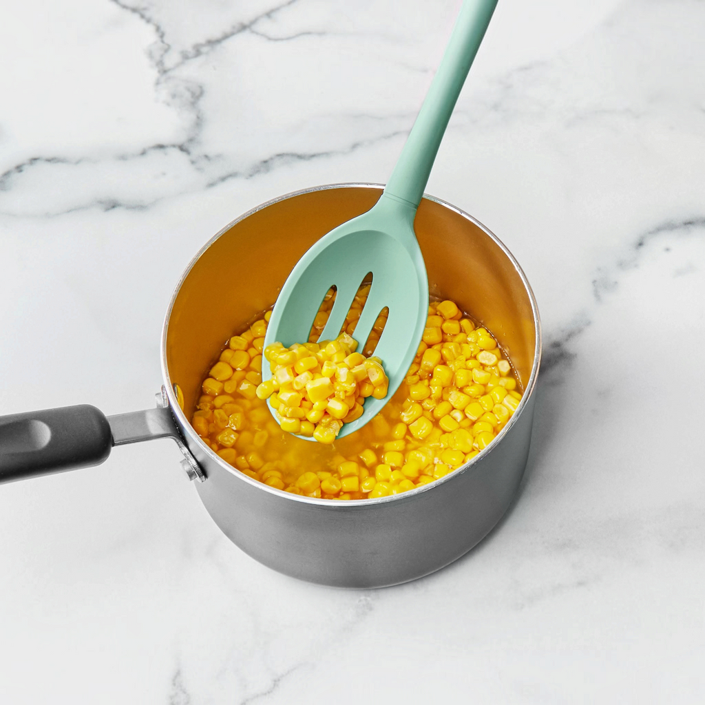 A saucepan with cooked corn kernels rests on marble as a tovolo Flex-Core Silicone Slotted Deep Spoon, in teal, lifts some corn from the pot.
