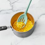 A saucepan with cooked corn kernels rests on marble as a tovolo Flex-Core Silicone Slotted Deep Spoon, in teal, lifts some corn from the pot.