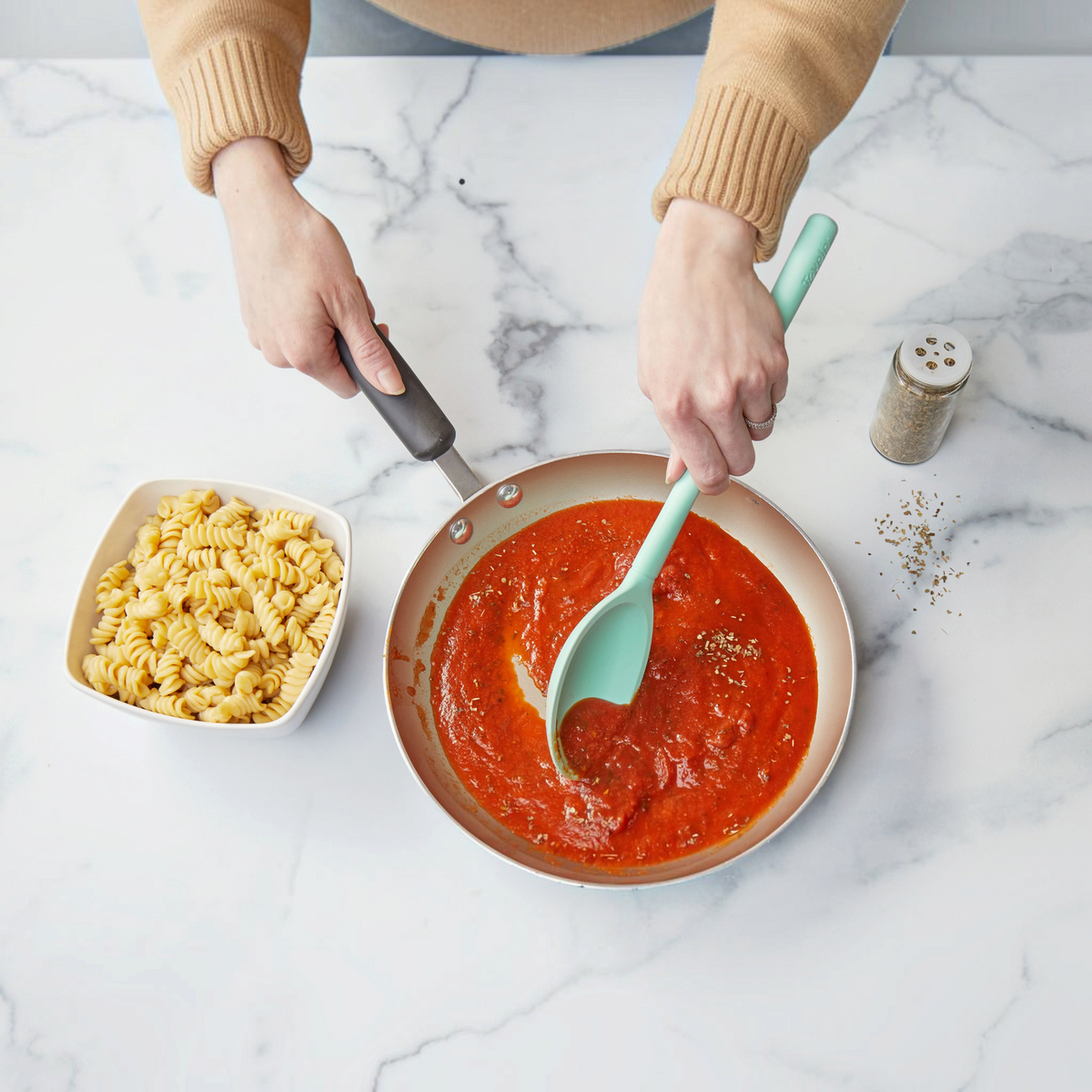 Someone stirs tomato sauce in a frying pan using the tovolo Flex-Core Silicone Deep Spoon beside a bowl of cooked rotini pasta on a marble countertop, with a jar of seasoning nearby.