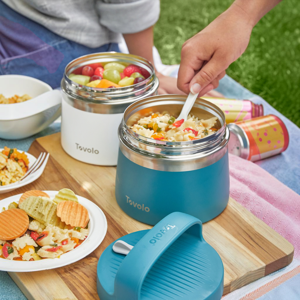 A hand scoops pasta salad from a blue Insulated Food Traveler container on a picnic table. Nearby, a white Tovolo container holds mixed fruit, with a plate of chips beside them.