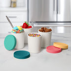 Four Mini Sweet Treats Tubs with colorful silicone lids sit on a modern kitchen counter; three are filled with ice cream and topped with rainbow sprinkles, granola, and strawberries. These tubs help prevent freezer burn.