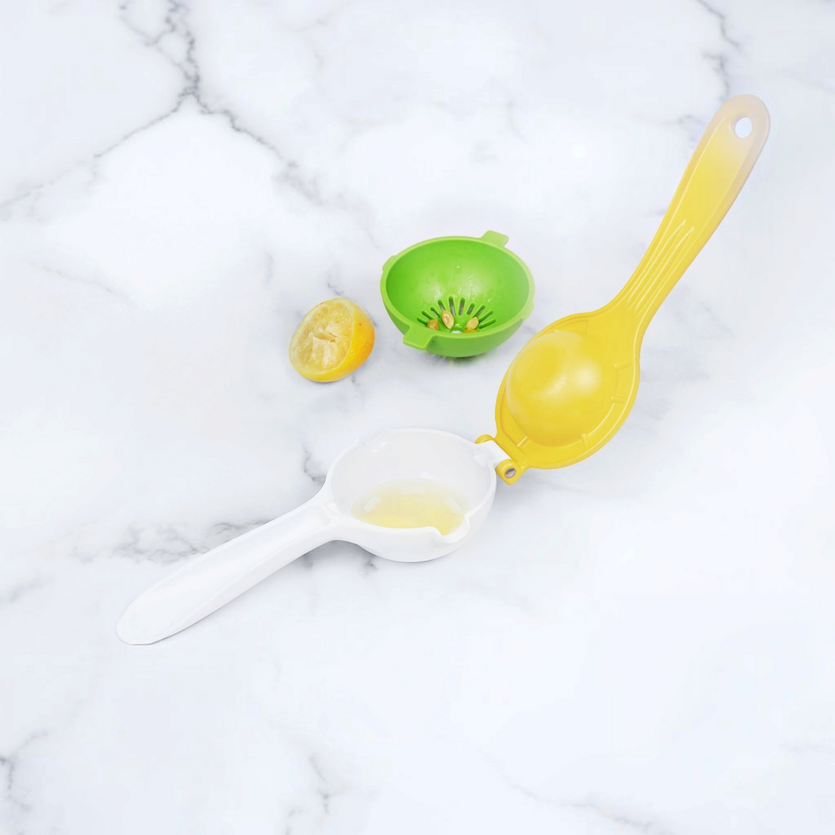 The Squeeze and Pour Citrus Press, featuring a yellow and white mess-free design, sits open next to a green juicer, a squeezed lemon half, and lemon seeds on a white marble surface.