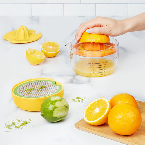 A hand squeezes an orange on the All-in-One Juicer filled with juice. Nearby are halved lemons, a zester with lime zest, whole oranges, a lime, and a wooden cutting board on a white kitchen counter.