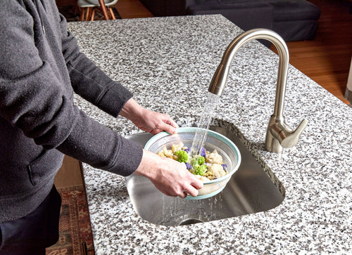 A person rinses mixed vegetables in a modern kitchen, preparing to cover them with the Collapsible Microwave Food Cover—made from high heat-resistant plastic—before microwaving.