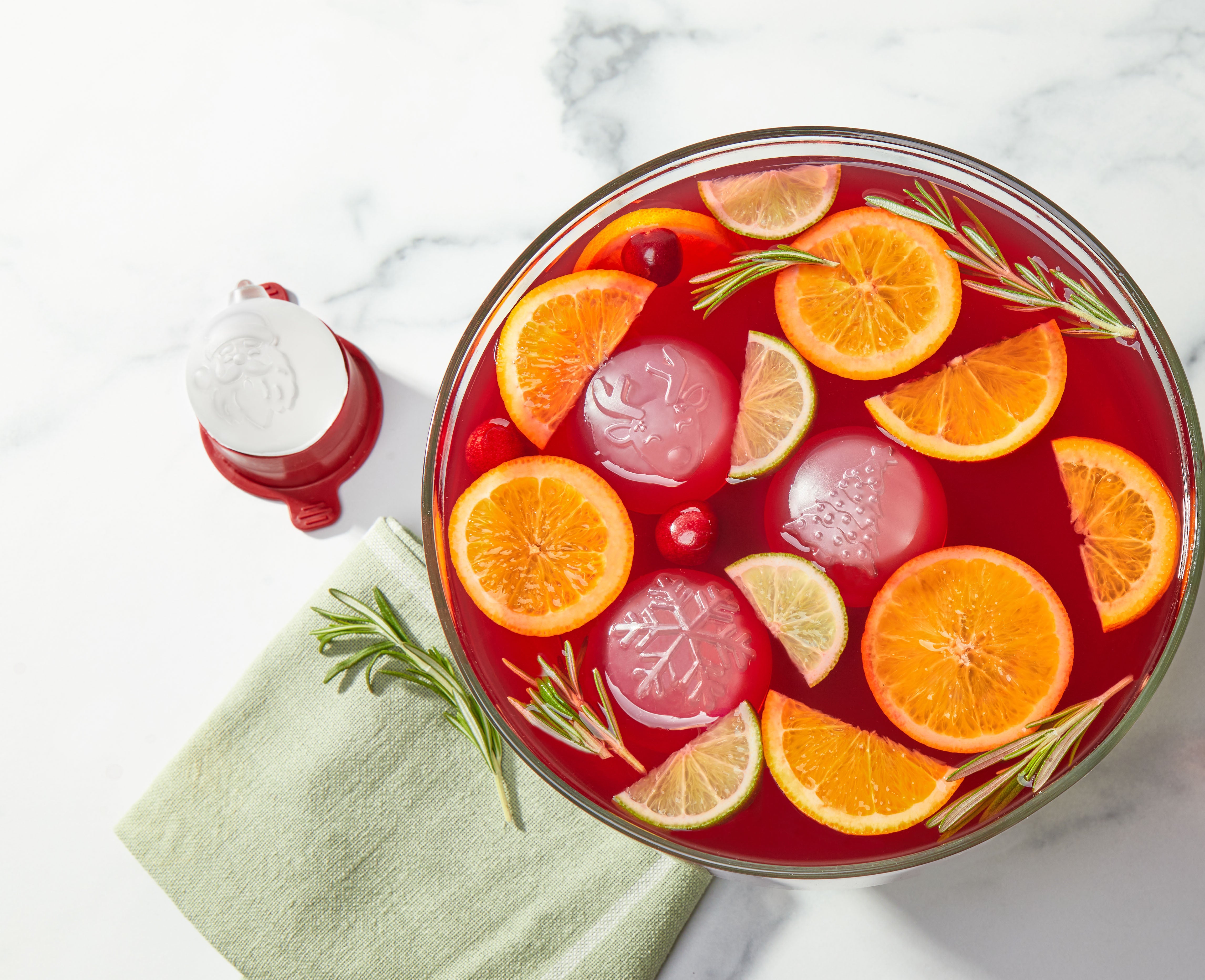 A bowl of red punch with orange and lime slices, cranberries, rosemary, and festive cocktail ice made using the Holiday Ice Mold sits on a marble surface beside a green napkin and Santa-shaped container.