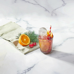 A glass of pink iced drink with orange slices, a cherry, and striped straw sits beside festive tovolo Santa Paws Ice Mold (Set of 2), an orange half, cherries, and a pine branch on a white surface.