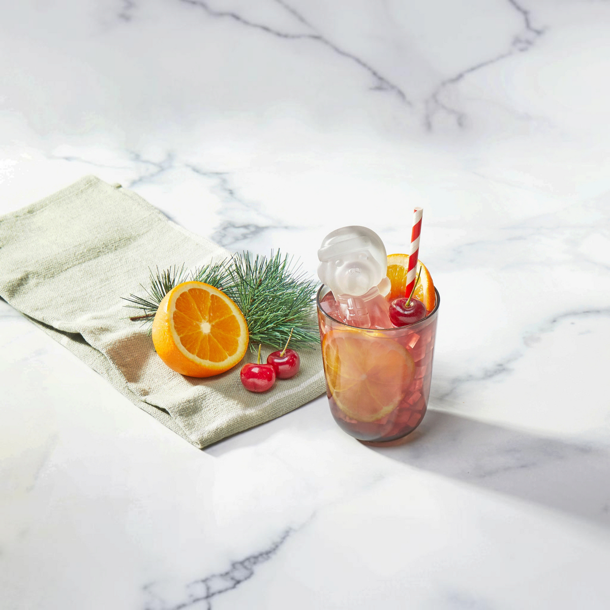 A glass of pink iced drink with orange slices, a cherry, and striped straw sits beside festive tovolo Santa Paws Ice Mold (Set of 2), an orange half, cherries, and a pine branch on a white surface.