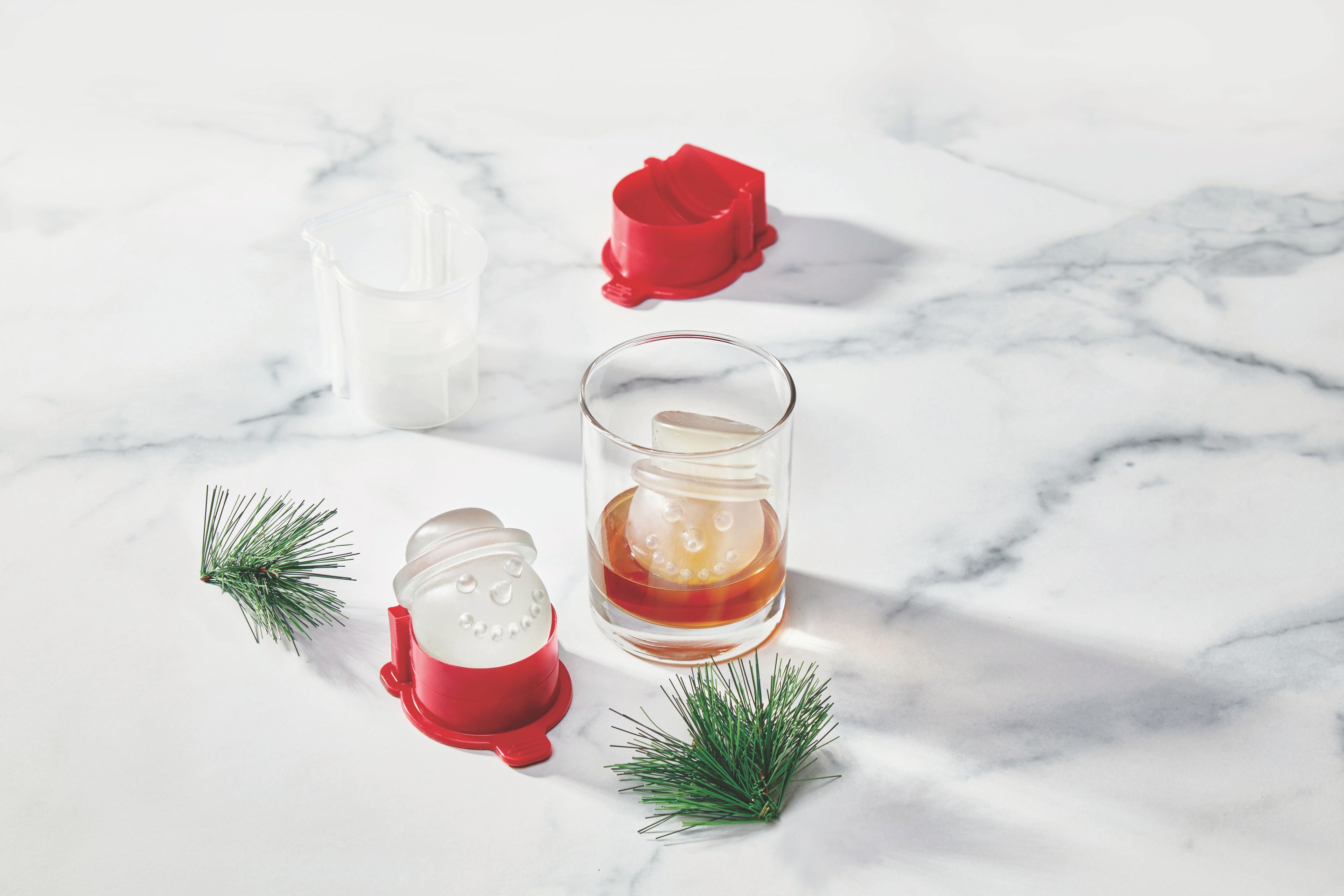 A glass of brown liquid with a snowman-shaped ice cube sits on a marble surface. Nearby are an empty snowman ice mold, a red ice mold base, and two small artificial pine branches.