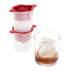A glass of dark liquid with a slow-melting, snowman-shaped ice cube made using tovolo's Top Hat Snowman Ice Molds (Set of 2) sits in front of two stacked red and white molds.