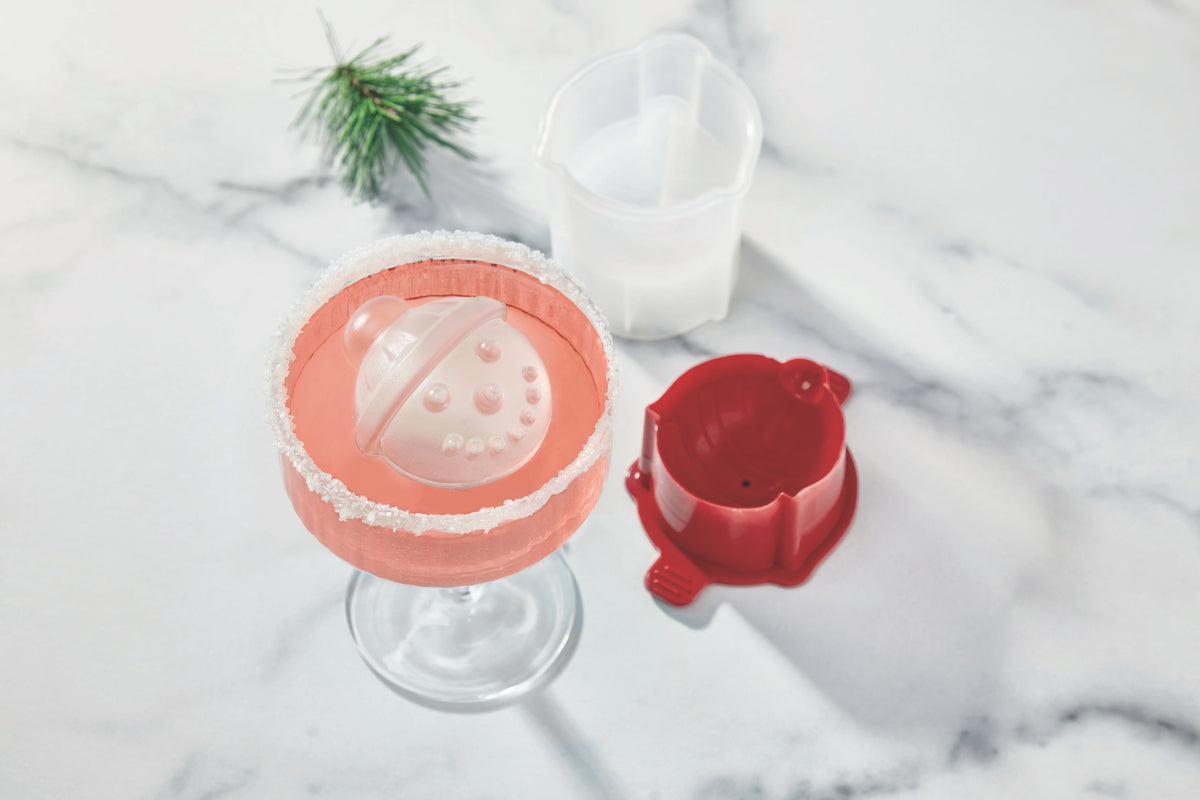 A pink cocktail with a salted rim sits on marble, featuring a large spherical ice cube made with tovolo’s Snowman Ice Molds (Set of 4). A pine sprig and white measuring cup add festive flair—perfect for holiday gatherings or Christmas gifting.