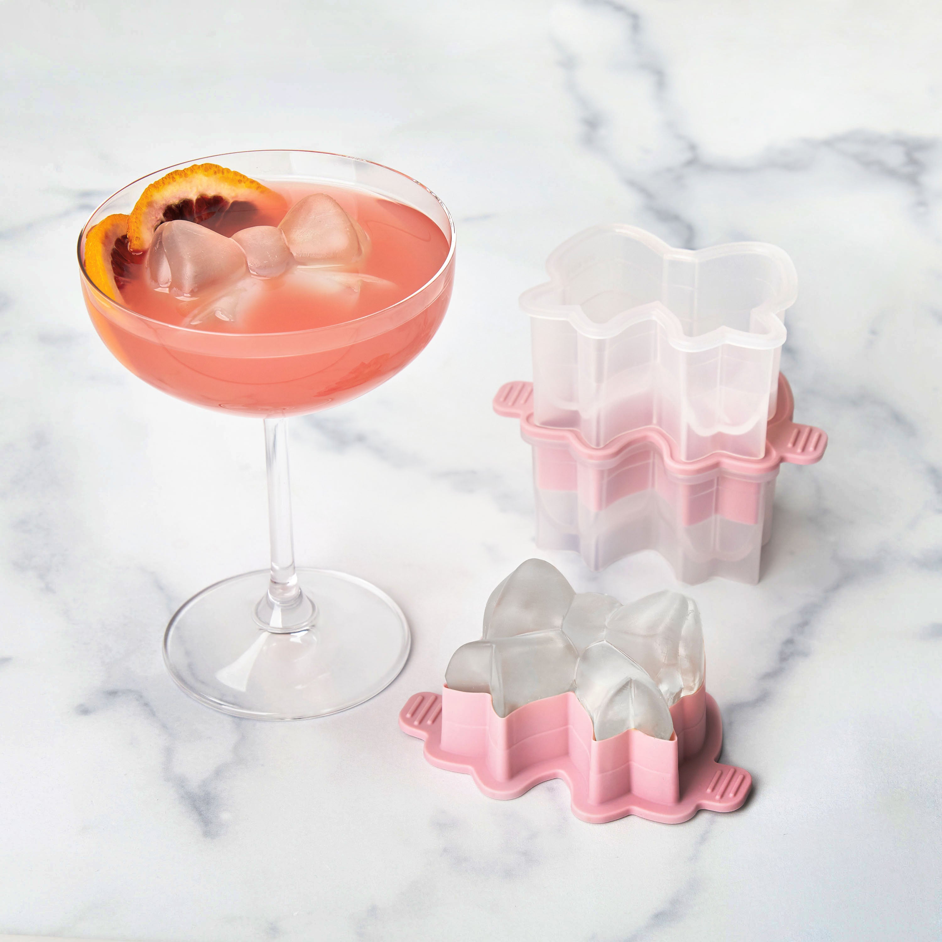 A pink cocktail with a bow-shaped ice cube and orange slice sits beside the tovolo Fancy Bow Ice Mold (Set of 2), in pink and white silicone, on a white marble surface.