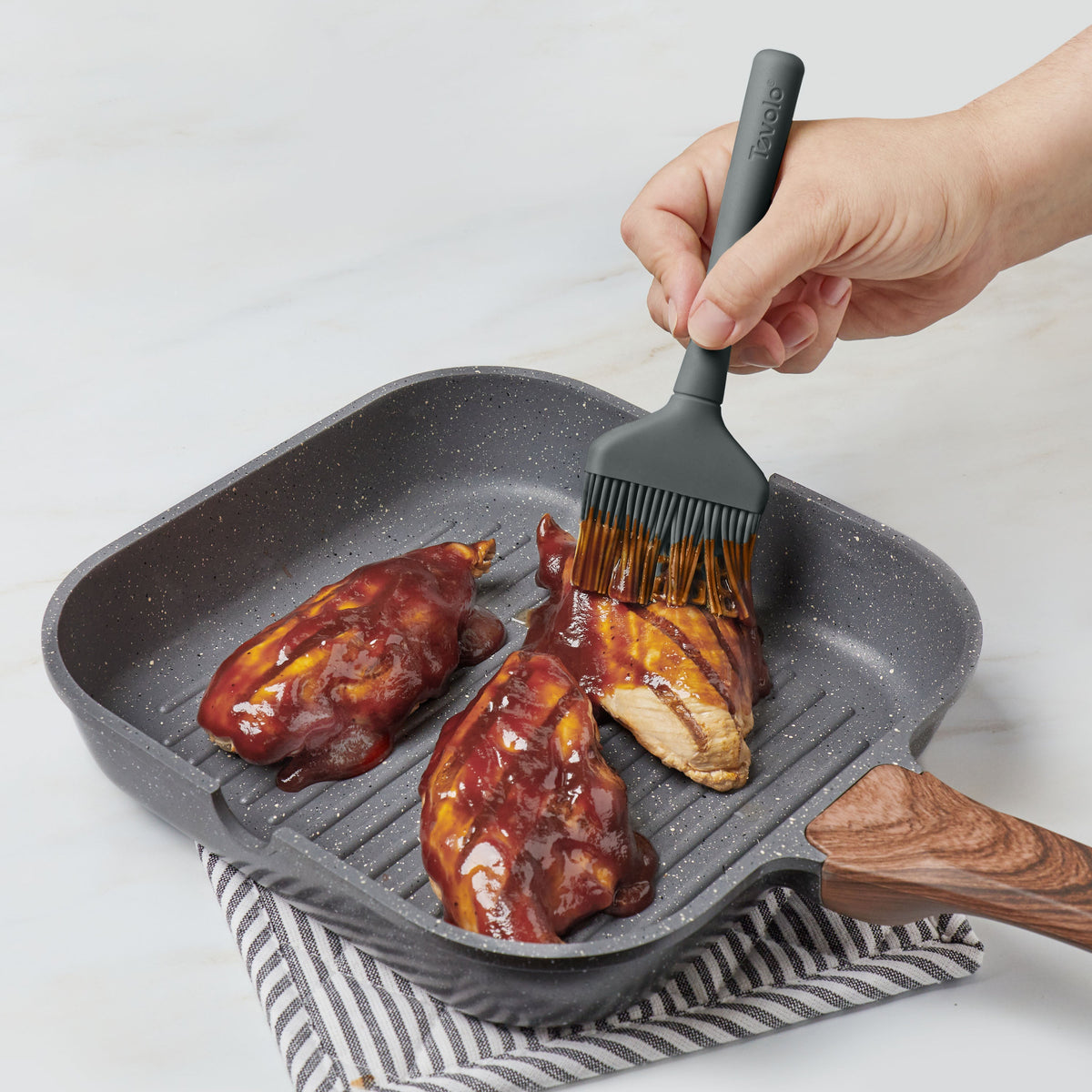 A hand uses the Flex-Core Basting Brush to spread barbecue sauce on three pieces of chicken cooking in a grill pan, which sits on a striped cloth atop a white surface.