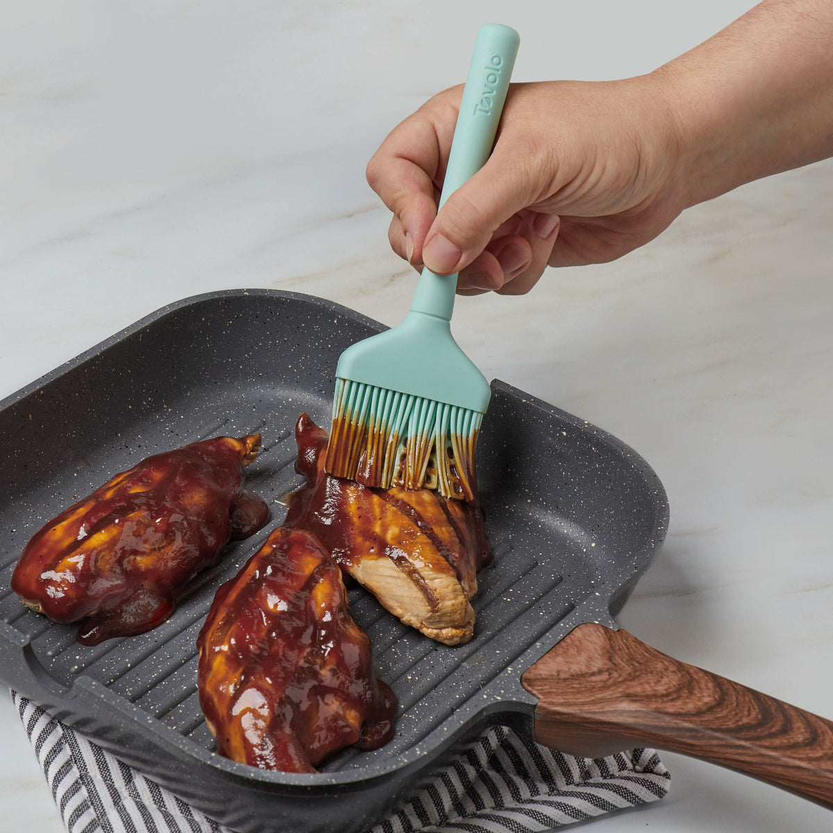 A hand uses the Flex-Core Basting Brush to spread barbecue sauce on three pieces of chicken cooking in a grill pan with a wooden handle, set on a striped cloth.
