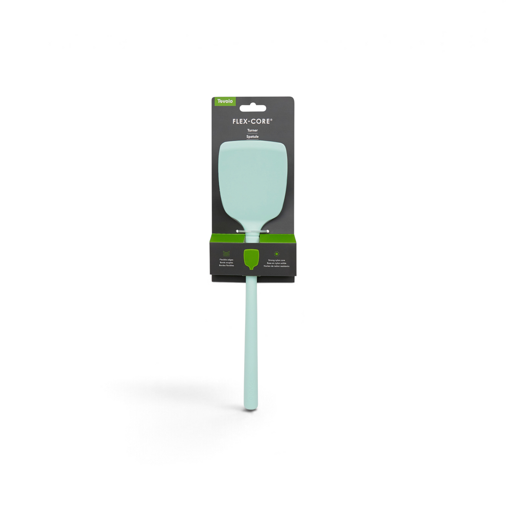 A tovolo Flex-Core Silicone Solid Turner in light green is shown in its packaging, which features a wide flexible head, long handle, and black packaging with green accents and white text.