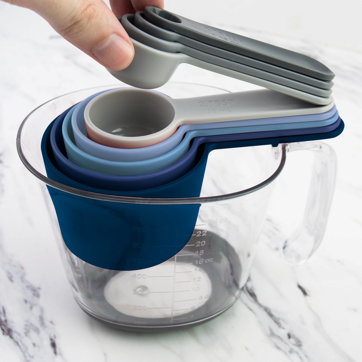 A hand lifts a gray etched spoon from the Magnetic Nested Measuring System, featuring colorful cups and spoons stacked inside a clear glass measuring cup on a white marble surface.