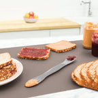 On the kitchen counter are jars of peanut butter and jelly, whole grain bread with spreads, a sandwich on a plate, and the Scoop and Spread kitchen tool. A bowl of fruit sits in the background.