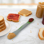 The Scoop and Spread, a double-ended kitchen tool with peanut butter on one end and jelly on the other, rests on a marble counter with bread slices, jars, and sandwiches—ideal for easy scooping and spreading.