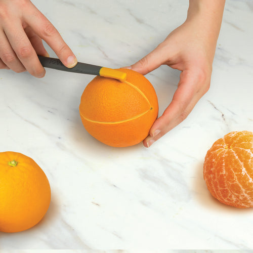 Using the 2-in-1 Slice and Peel Citrus Tool, a person cuts a line around the middle of an orange on a marble surface, with the tool, a whole orange, and a peeled orange nearby.