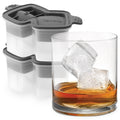 A glass of whiskey with two large, slow-melting cubes sits by stacked tovolo Double Rocks Ice Molds (Set of 2), featuring gray and white silicone with geometric lid designs.