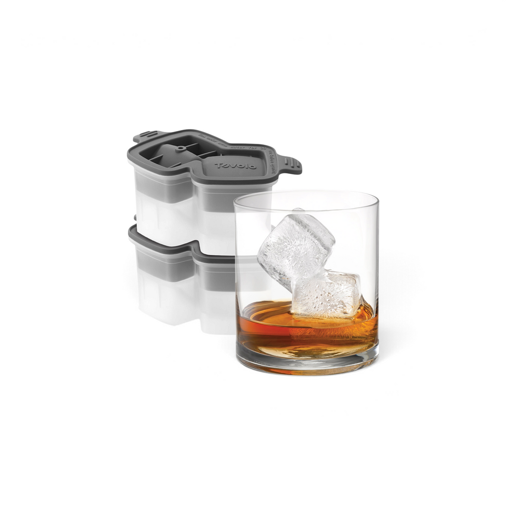A glass of whiskey with two large, slow-melting cubes sits by stacked tovolo Double Rocks Ice Molds (Set of 2), featuring gray and white silicone with geometric lid designs.