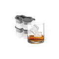 A glass of whiskey with two large, slow-melting cubes sits by stacked tovolo Double Rocks Ice Molds (Set of 2), featuring gray and white silicone with geometric lid designs.