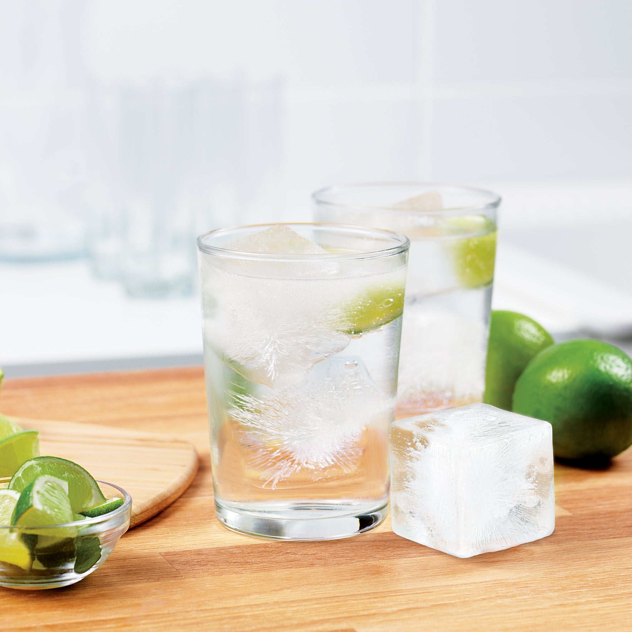 Two glasses of sparkling water with extra-large ice from the tovolo King Cube Silicone Ice Molds (Set of 2) and fresh lime wedges sit on a wooden surface, with additional limes and blurred glassware in the background.