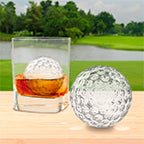 A glass of whiskey with a baseball-shaped ice cube made by tovolo’s Baseball Ice Molds (Set of 2) sits on a wooden surface outdoors, another identical ice cube beside it, with grass, trees, and a small lake in the background.