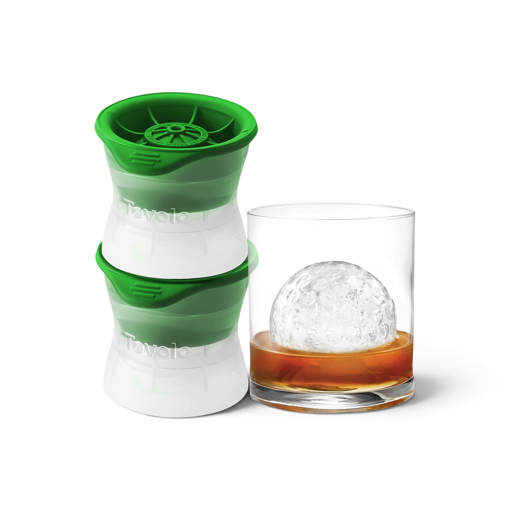 Two green and white Tovolo Baseball Ice Molds (Set of 2) are placed beside a glass of whiskey, displaying a large, clear baseball-shaped ice sphere inside.