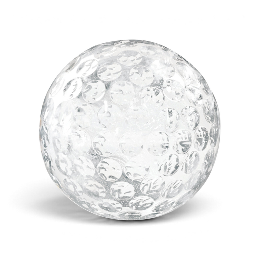 A clear, round ice sphere shaped like a baseball, made with tovolo Baseball Ice Molds (Set of 2), rests on a white surface with a subtle shadow underneath.