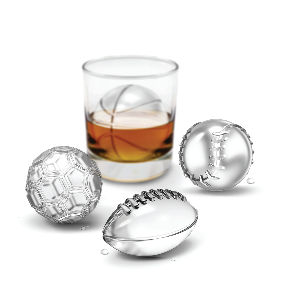 Three metal balls—shaped like a soccer ball, football, and baseball—sit next to a whiskey glass holding a basketball ice ball, highlighting our Sports Ice Molds for the perfect sports-themed chill.