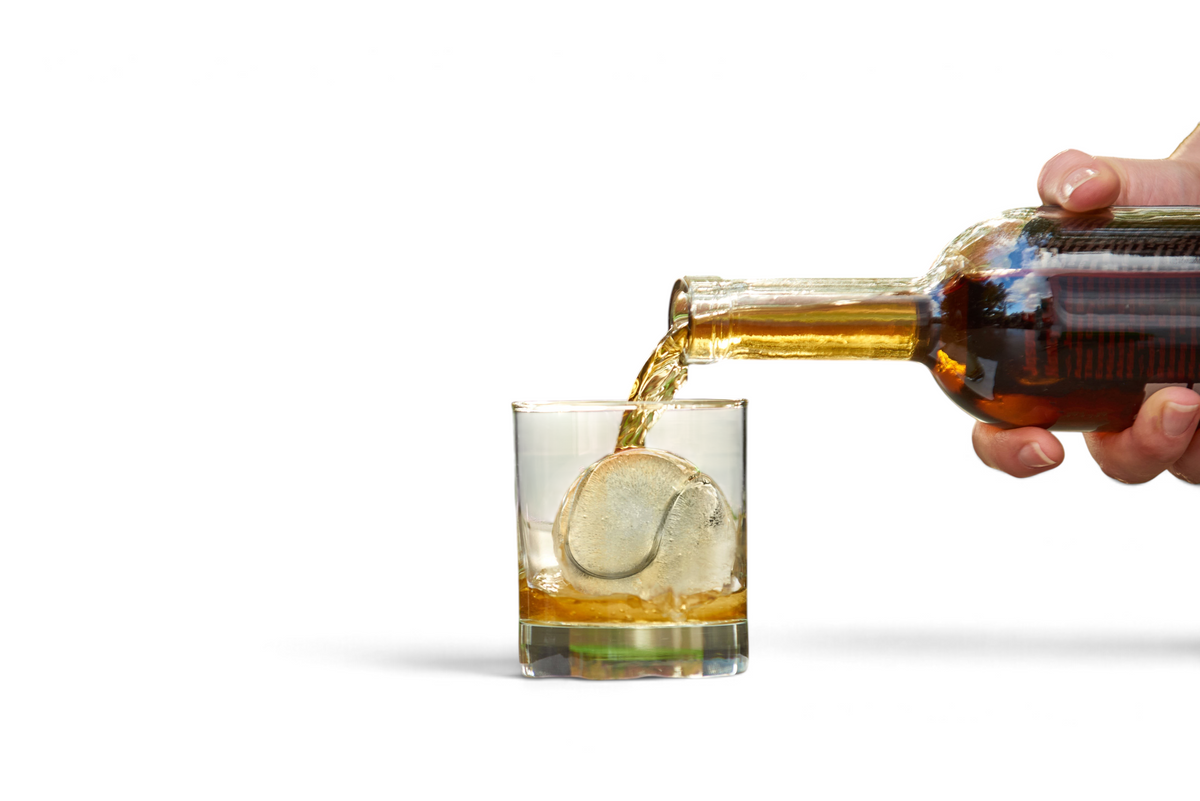 A hand pours amber liquid from a bottle into a glass with a large round ice cube made using tovolo Baseball Ice Molds (Set of 2), outdoors on a table with a blurred green and brown background.