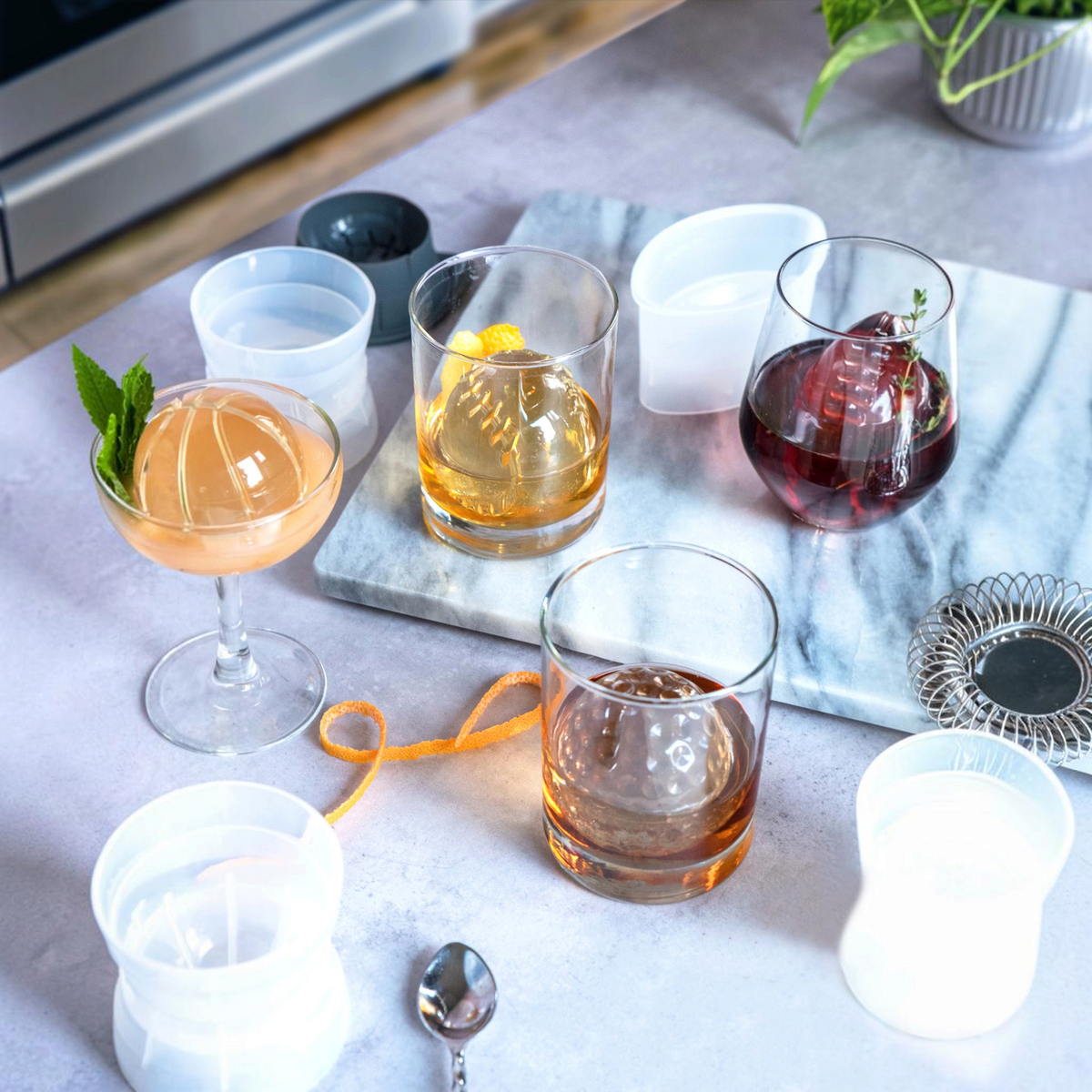 A marble tray displays three cocktails with large baseball-shaped ice cubes made using tovolo Baseball Ice Molds (Set of 2) in assorted glasses. Nearby are empty stackable molds, a spoon, orange peel, and a spring strainer on a light surface.