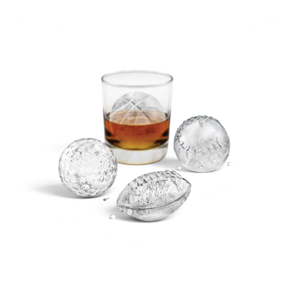A glass of whiskey with a spherical ice cube sits in the background, while tovolo Baseball Ice Molds (Set of 2) create golf ball, football, and baseball-shaped cubes on a white surface dotted with water droplets.