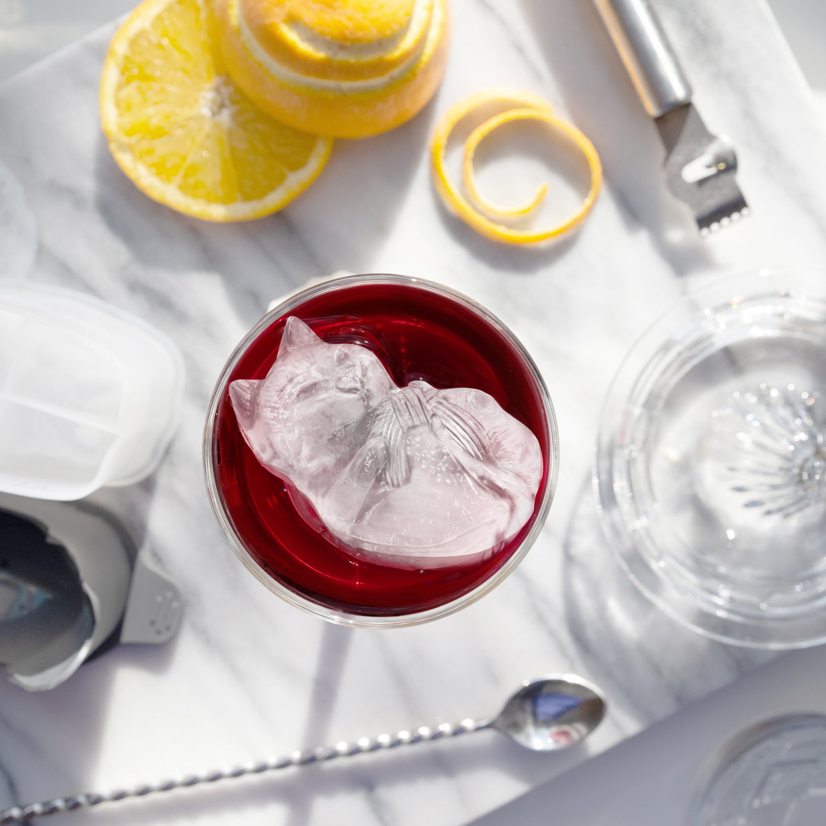 A glass of red drink with a dog-shaped ice cube, made using tovolo Dog Ice Molds (Set of 2), sits on marble beside sliced lemon, lemon peel, a zester, metal spoon, and glass citrus juicer.