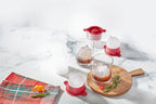 Two glasses with round ice made from tovolo Festive Holiday Ornament Ice Molds (Set of 4) sit on a wooden board, surrounded by the molds, orange slices, rosemary, and a plaid napkin on a white marble surface.