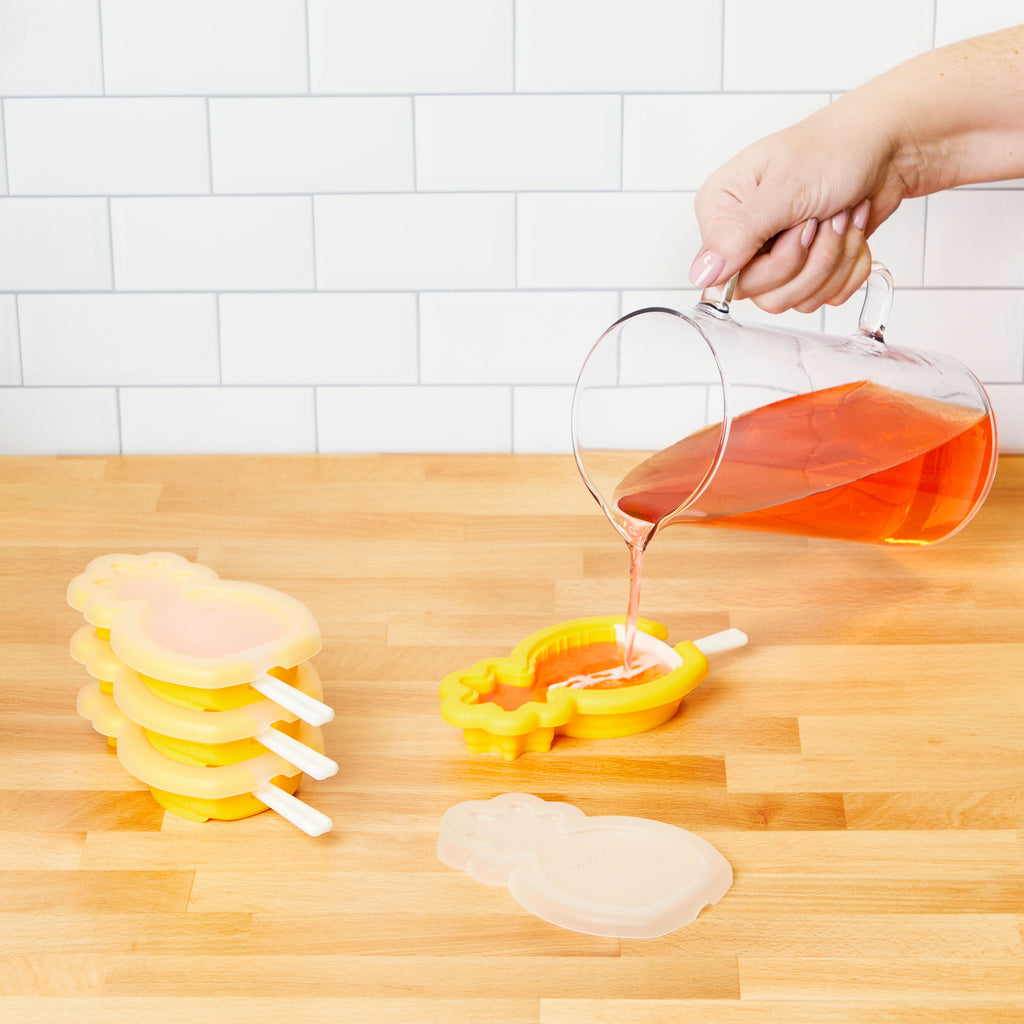 A hand pours orange liquid from a glass pitcher into a yellow pineapple-shaped Stackable Pop Mold by tovolo on a wooden countertop, with more popsicle molds and a white tiled wall in the background.