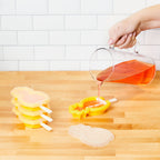 A hand pours orange liquid from a glass pitcher into a yellow pineapple-shaped Stackable Pop Mold by tovolo on a wooden countertop, with more popsicle molds and a white tiled wall in the background.