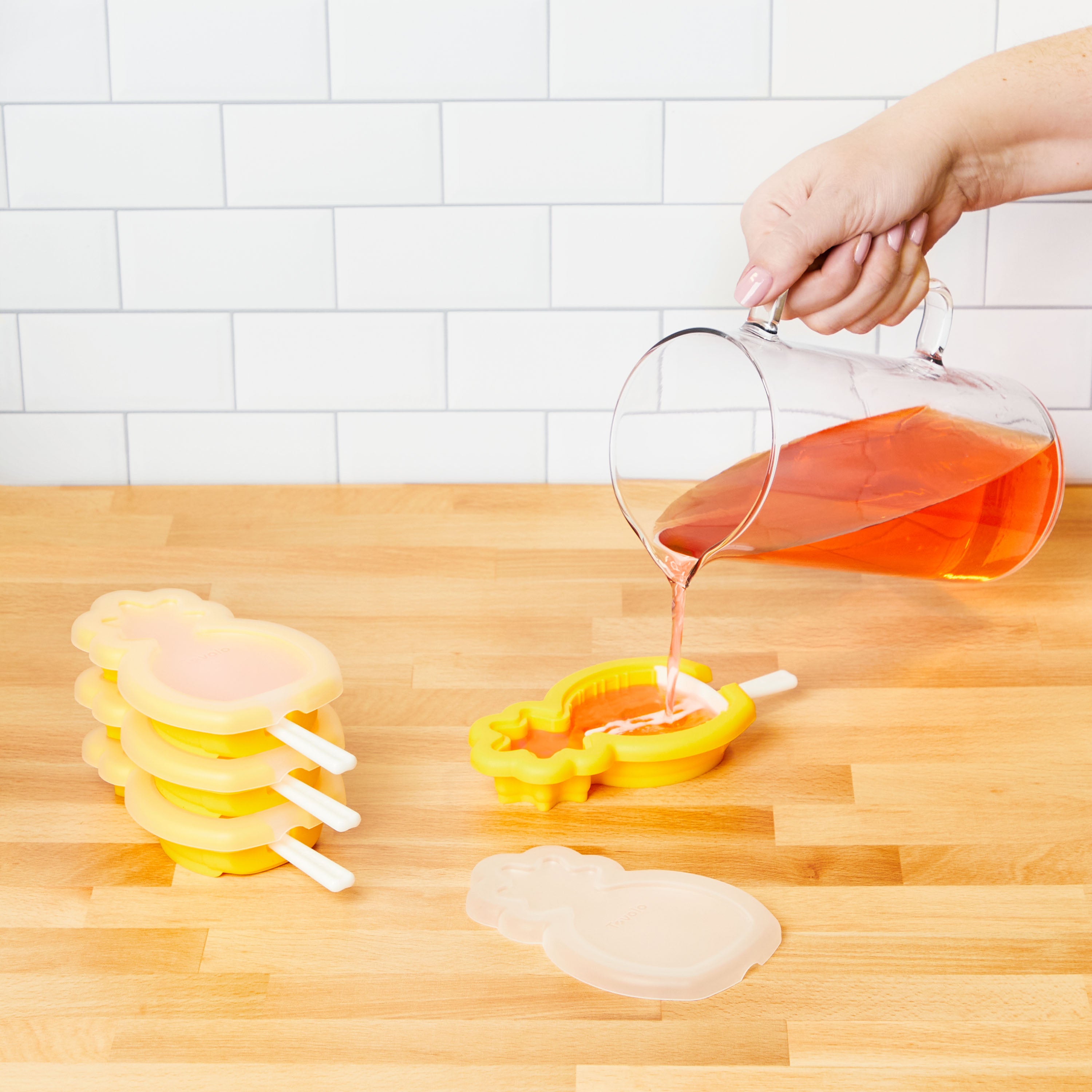 A hand pours orange liquid from a glass pitcher into a yellow pineapple-shaped Stackable Pop Mold by tovolo on a wooden countertop, with more popsicle molds and a white tiled wall in the background.