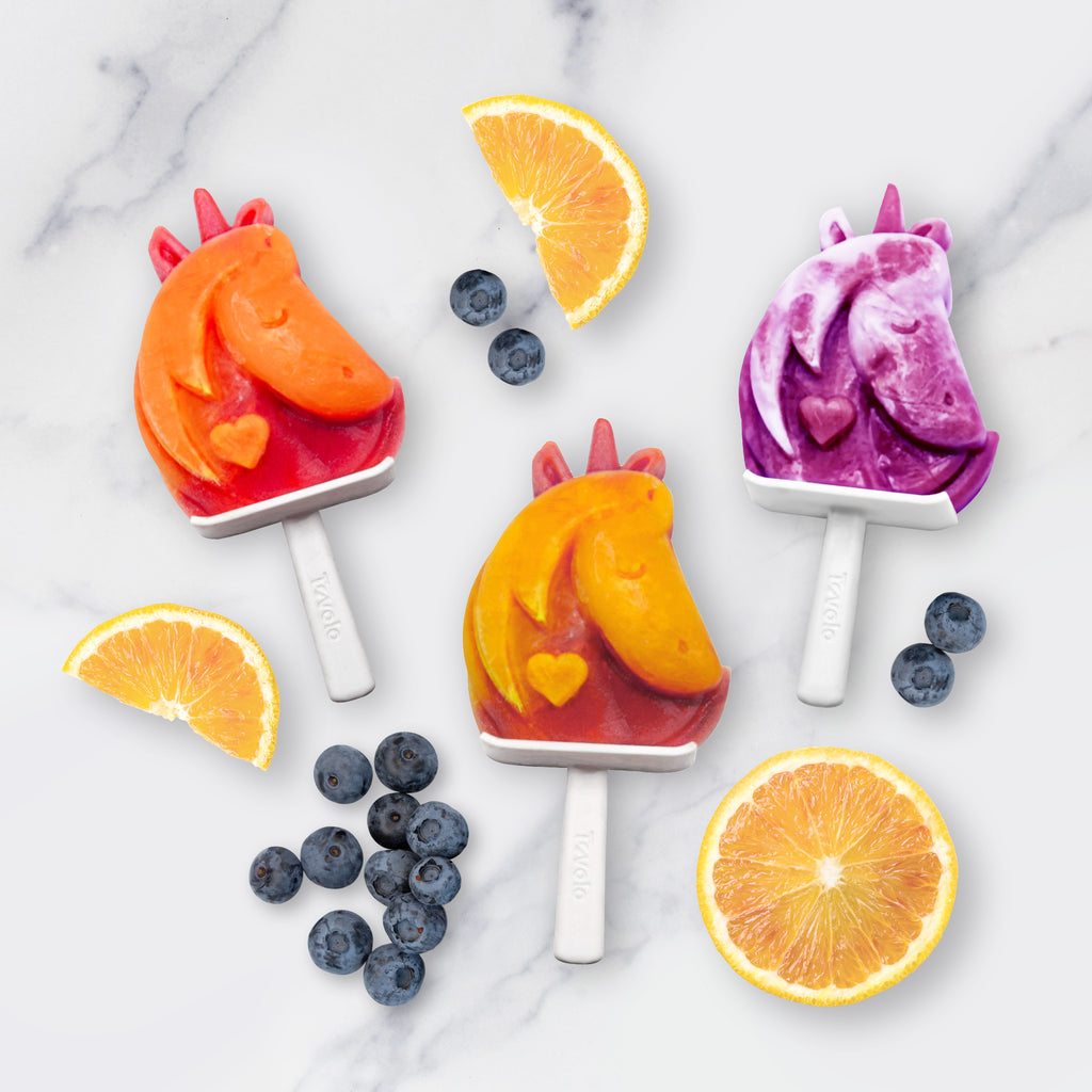 Three unicorn-shaped popsicles—red, orange, and purple—sit on a marble surface with blueberries and orange slices, made using Stackable Pop Molds for easy homemade treats.